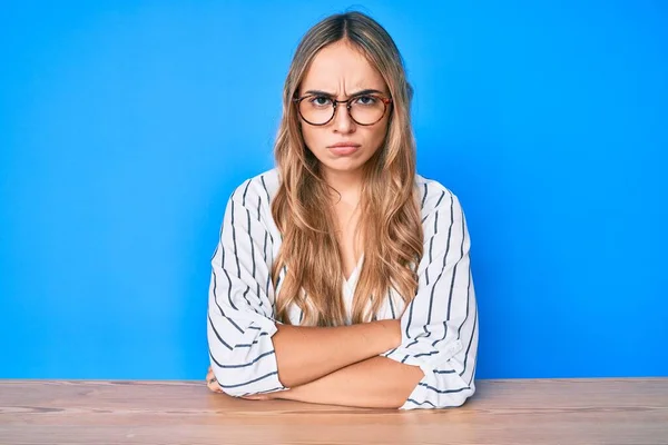 若い美しいブロンドの女性が眼鏡をテーブルの上に座って懐疑的で緊張し 両腕を組んで顔に不承認の表現 否定的な人 — ストック写真