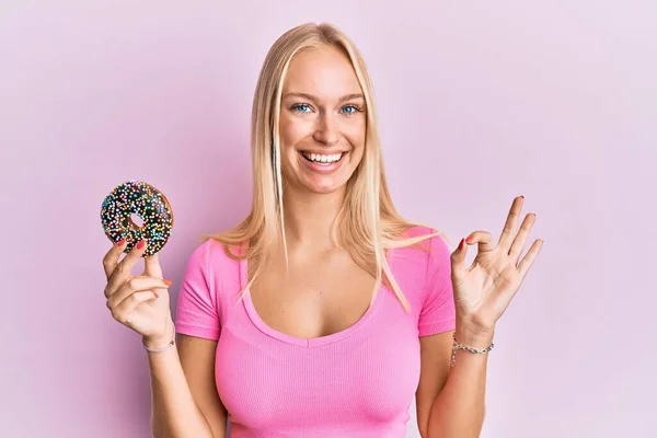 Menina Loira Jovem Segurando Donut Fazendo Sinal Com Dedos Sorrindo — Fotografia de Stock