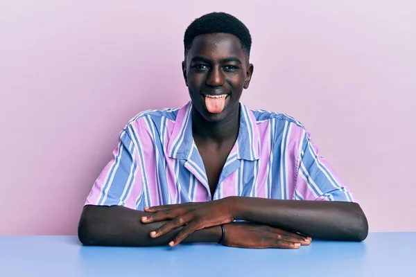 身穿休闲装的年轻的非洲裔美国人坐在桌子上 带着滑稽的表情伸出舌头 情感概念 — 图库照片