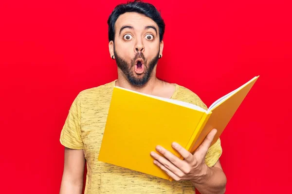 Young Hispanic Man Holding Book Scared Amazed Open Mouth Surprise —  Fotos de Stock