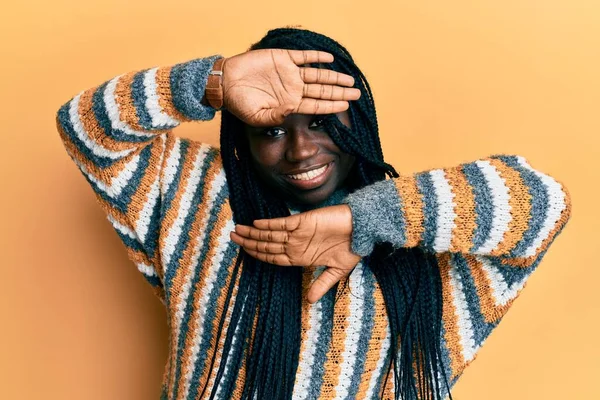 Young black woman with braids wearing casual winter sweater smiling cheerful playing peek a boo with hands showing face. surprised and exited