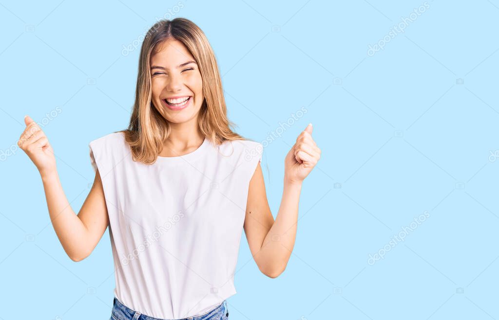 Beautiful caucasian woman with blonde hair wearing casual white tshirt very happy and excited doing winner gesture with arms raised, smiling and screaming for success. celebration concept. 