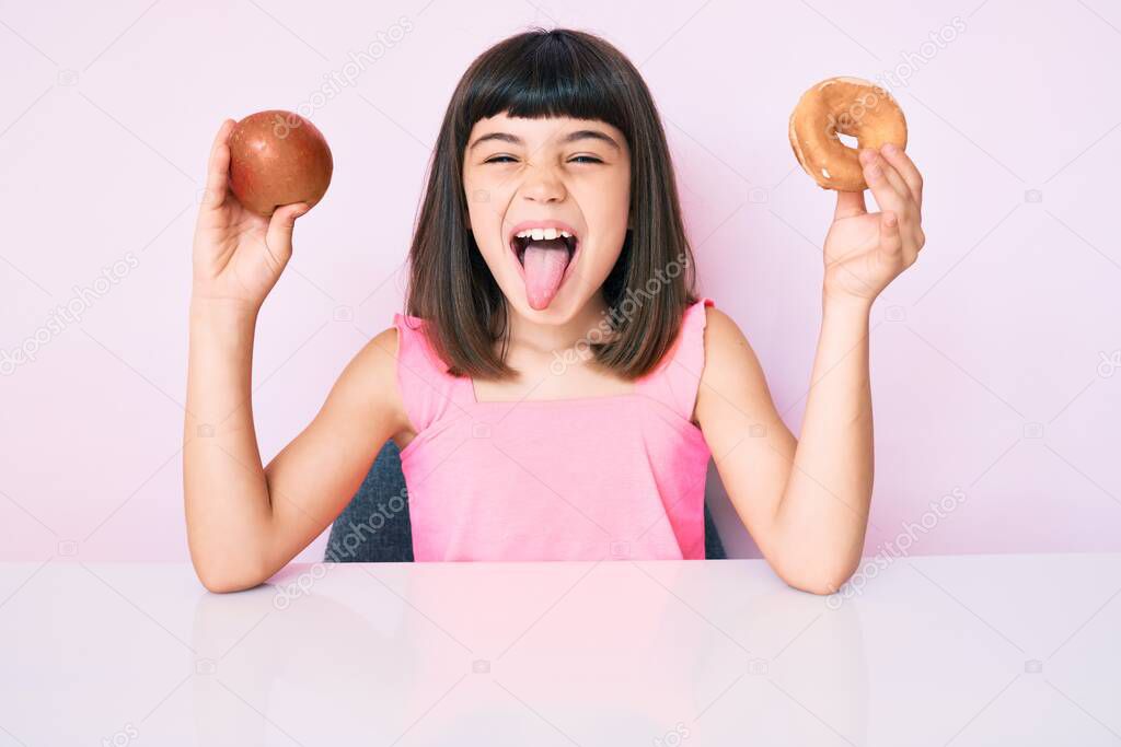 Young little girl with bang holding red apple and donut sitting on the table sticking tongue out happy with funny expression. 