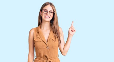 Genç, güzel, sarışın bir kadın. Günlük kıyafetler ve gözlük takıyor. Yüzünde kocaman bir gülümseme var.. 