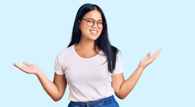 Genç ve güzel Asyalı kız günlük kıyafetler ve gözlük takıyor gülümsüyor iki elini de açık tutuyor, sunuyor ve reklam karşılaştırması yapıyor ve dengeyi sağlıyor. 