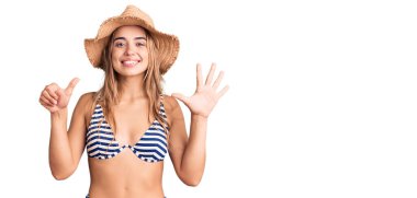 Bikini giyen, şapka takan, 6 numaralı parmaklarıyla işaret eden güzel, sarışın bir kadın. Kendinden emin ve mutlu gülümserken.. 