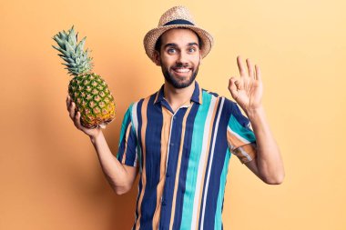 Young handsome man with beard wearing summer hat and shirt holding pineapple doing ok sign with fingers, smiling friendly gesturing excellent symbol 