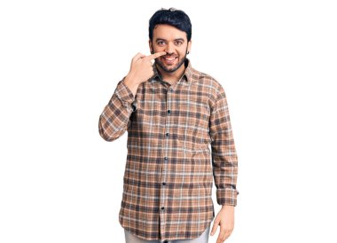 Latin asıllı genç bir adam, gündelik giysiler giyip el ve burnu işaret ediyor, neşeli bir şekilde gülümsüyor. güzellik konsepti 