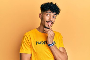 Mutluluk mesajı içeren tişört giyen genç Arap yakışıklı adam kameraya kollarını kavuşturup çenesine dokunarak gülümsüyor. Olumlu düşünmek. 