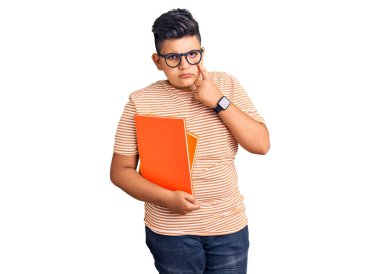 Little boy kid holding book wearing glasses pointing to the eye watching you gesture, suspicious expression 
