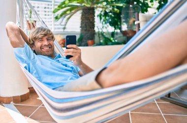 Orta yaşlı yakışıklı bir adam evinin terasında, bir hamakta telefonla dinleniyor.