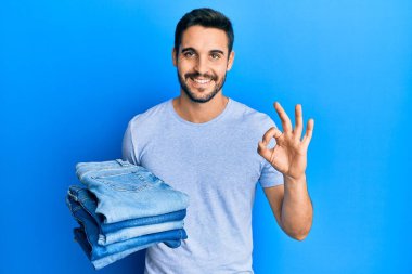 Bir yığın katlanmış kot tutan genç İspanyol adam parmaklarıyla imza atıyor, gülümseyerek mükemmel bir sembol oluşturuyor. 