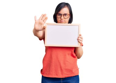 Down sendromlu esmer kadın elinde boş beyaz karatahta ile elinde dur işareti ile ciddi ve kendinden emin bir ifadeyle savunma hareketi yapıyor. 