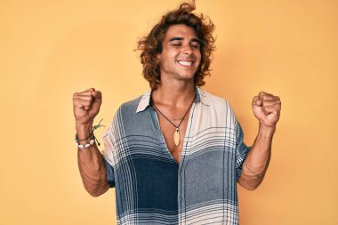 İspanyol asıllı genç adam yaz stili giyiniyor. Çok mutlu ve heyecanlı. Kolları havada, gülümseyen ve başarı için çığlık atan kazanan bir jest yapıyor. kutlama konsepti. 