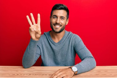 Yakışıklı İspanyol bir adam, sıradan giyinmiş masanın üzerinde oturuyor ve üç numaralı parmaklarıyla işaret ediyor kendinden emin ve mutlu gülümsüyor.. 