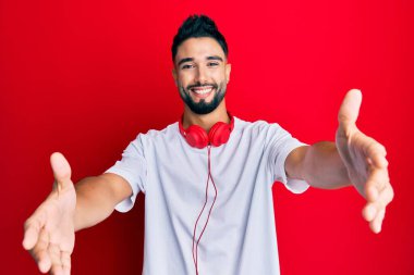 Sakallı genç adam müzik dinliyor kulaklık takıyor kameraya bakıyor kucaklaşmak için kollarını açmış gülümsüyor. Mutluluğu kucaklayan neşeli bir ifade.. 