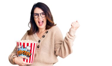 Beautiful young brunette woman eating popcorn screaming proud, celebrating victory and success very excited with raised arms 