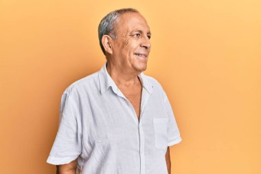 Yakışıklı, olgun, günlük beyaz tişört giyen, yüzünde gülümseme ve doğal ifadeyle yan yana bakan bir adam. Kendinden emin gülen. 