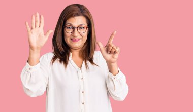 Orta yaşlı Latin bir kadın günlük kıyafetler ve gözlük takıyor. Yedi numaralı parmaklarıyla işaret ediyor ve gülümsüyor. Kendinden emin ve mutlu.. 