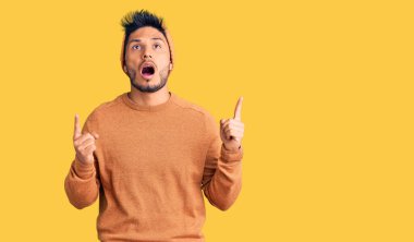 Yakışıklı Latin Amerikalı süveter ve yün şapka giyen genç adam şaşırmış ve şaşırmış bir şekilde yukarı bakıyor ve parmaklarıyla işaret ediyor ve kollarını kaldırıyordu.. 