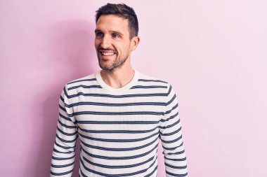 Genç, çizgili süveter giyen yakışıklı bir adam soyutlanmış pembe arka planda dikilip yan yana bakıyor, rahat profil pozu veriyor doğal bir yüzü ve kendine güvenen bir gülümsemesi var..