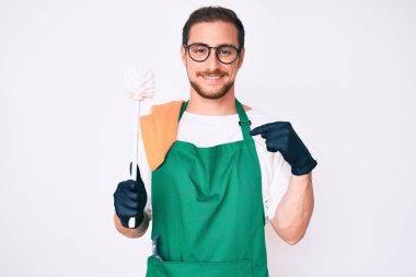 Önlük takmış genç yakışıklı bir adam tuvalet fırçasını tutuyor mutlu ve gururlu gülümseyen birini işaret ediyor. 