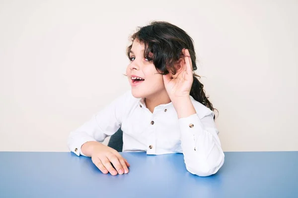 耳を傾け 噂やゴシップに耳を傾け手で笑みを浮かべてテーブルの上に座ってカジュアルな服を着てかわいいヒスパニック系の子供 聴覚障害の概念 — ストック写真