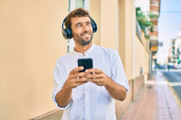 晴れた日に白いシャツを着て髭を生やしたハンサムな男は ヘッドフォンを着て音楽を聞いてスマートフォンを使って屋外で幸せな笑顔 — ストック写真