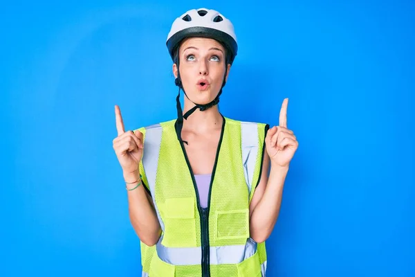 Young caucasian girl wearing bike helmet and reflective vest amazed and surprised looking up and pointing with fingers and raised arms.