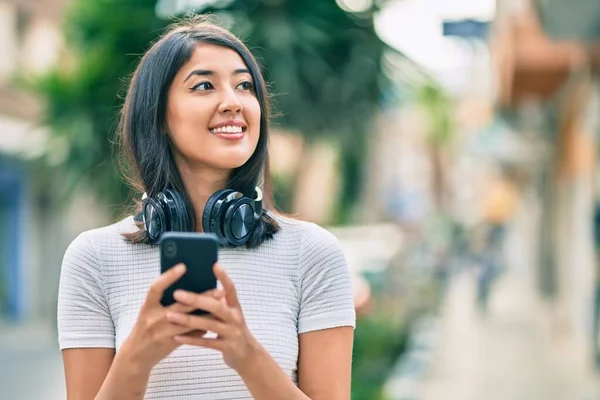 街中でスマホやヘッドフォンを使って笑顔で幸せそうな若いヒスパニック系女性 — ストック写真