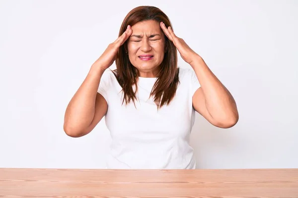 ストレスのため 頭の上に手でテーブルの上に座ってカジュアルな白いTシャツを着て中世のブルネットのヒスパニック系の女性 片頭痛 — ストック写真