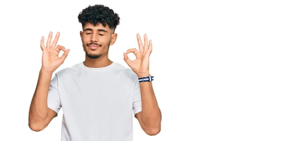 カジュアルな白いTシャツを着た若いアラブ人男性がリラックスして笑顔で目を閉じて指で瞑想のジェスチャーをしています ヨガのコンセプト — ストック写真