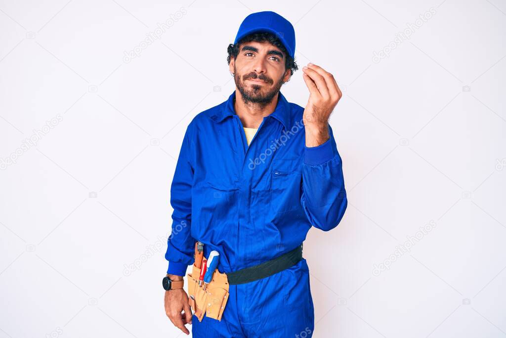 Handsome young man with curly hair and bear weaing handyman uniform doing italian gesture with hand and fingers confident expression 