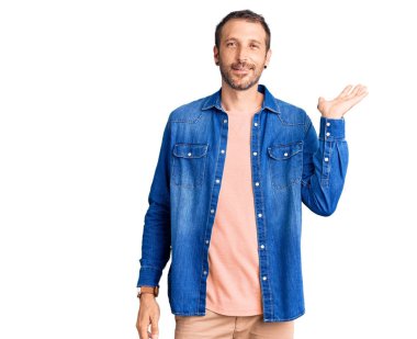 Gündelik kıyafetler giyen, neşeli gülümseyen, eli avucunda kameraya bakan genç ve yakışıklı bir adam.. 