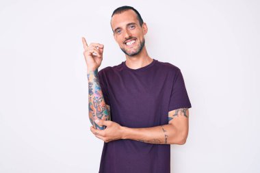 Dövmeli, sıradan kıyafetler giyen, yüzünde kocaman bir gülümseme olan, eli ve parmağı kameraya bakan genç yakışıklı bir adam.. 