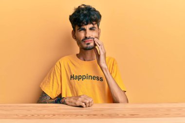 Üzerinde mutluluk mesajı olan tişört giyen genç İspanyol adam diş ağrısı ya da diş ağrısı yüzünden masaya oturmuş ağzıyla dokunuyor. dişçi 