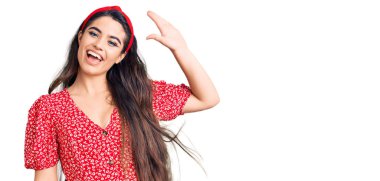 Yazlık elbise giyen esmer kız merhaba diyor mutlu ve gülümsüyor, dostça karşılama hareketi yapıyor. 