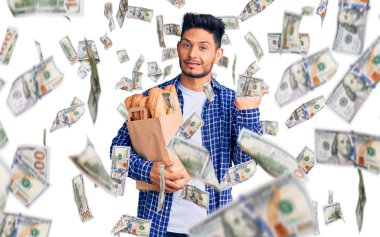 Yakışıklı Latin Amerikalı genç adam, elinde kağıt torbayla arkasını göstererek elini ve parmağını kaldırmış, kendinden emin gülümsüyor.