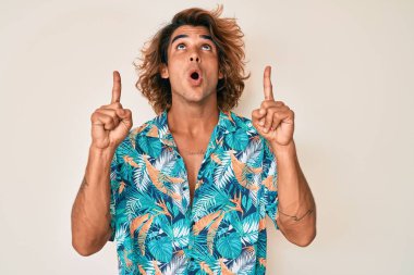 Yaz tişörtü giyen İspanyol genç adam şaşırmış ve şaşırmış bir şekilde yukarı bakıyor ve parmaklarıyla işaret ediyor ve kollarını kaldırıyor.. 