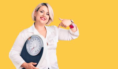 Genç sarışın, kilolu, elinde tartı makinesi olan bir kadın kendini mutlu ve gururlu bir şekilde gösteriyordu. 