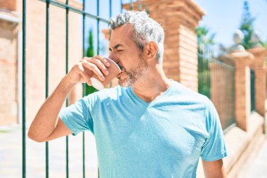 Orta yaşlı, kır saçlı, kahve içen, şehir caddesinde yürüyen bir adam..