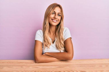 Güzel, sarışın, gündelik giysiler giyen mutlu yüzlü, kollarını kavuşturup kameraya bakan genç bir kadın. pozitif kişi. 