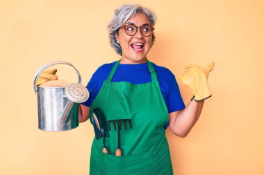 Bahçıvan önlüğü giymiş yaşlı, beyaz saçlı bir kadın ve elinde sulama kabı tutan eldivenler başparmağıyla yan tarafa doğru gülümsüyor ve ağzı açık gülümsüyor. 