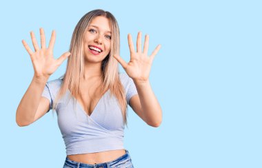 Genç, güzel, sarışın, gündelik giysiler giyen, on numaralı parmaklarıyla işaret eden, kendinden emin ve mutlu bir şekilde gülümseyen bir kadın.. 