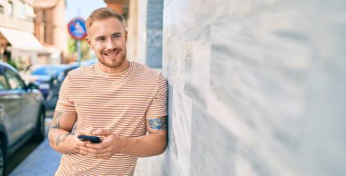 Genç İrlandalı mutlu bir şekilde gülümsüyor. Şehrin duvarına yaslanmış akıllı telefon kullanıyor..