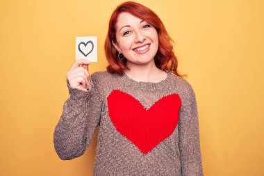 Genç, güzel, kızıl saçlı bir kadın, sarı arka plan üzerinde kalbi ile hatırlatıcı taşıyor. Pozitif ve mutlu görünüyor. Kendine güvenen bir gülümsemeyle dişlerini gösteriyor.