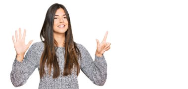 Genç esmer kadın günlük kışlık süveter giyer ve yedi numaralı parmaklarıyla işaret ederken kendinden emin ve mutlu bir şekilde gülümser.. 