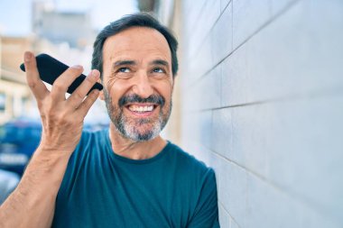 Orta yaşlı sakallı bir adam dışarıda mutlu bir şekilde gülümsüyor telefonda sesli mesaj dinliyor.