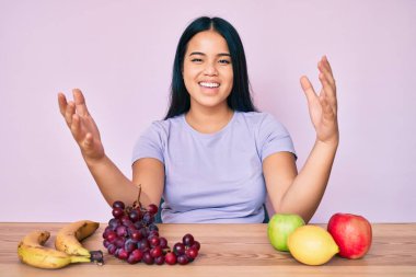 Genç ve güzel Asyalı kız taze ve sağlıklı meyve yiyor kameraya bakıyor kucaklaşmak için kollarını açmış gülümsüyor. Mutluluğu kucaklayan neşeli bir ifade.. 