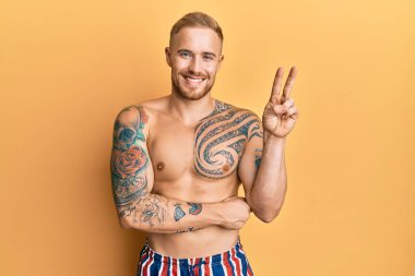 Mayo giyen genç, beyaz bir adam. Yüzü gülüyor, kameraya göz kırpıyor ve zafer işareti yapıyor. İki numara.. 